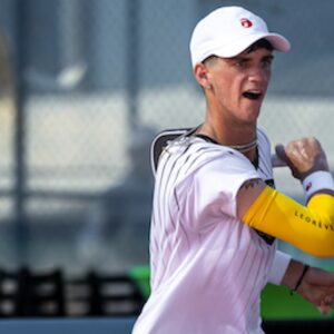 A pickleball player in a white shirt and black shorts is preparing to hit a shot on an outdoor court. He is wearing a white cap and a yellow sleeve on his left arm.