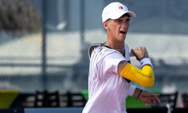 A pickleball player in a white shirt and black shorts is preparing to hit a shot on an outdoor court. He is wearing a white cap and a yellow sleeve on his left arm.